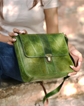 A woman holds a green real leather shoulder bag with a front flap and gold-tone clasp, set against a blurred outdoor background. 100% genuine full grain leather, natural patina