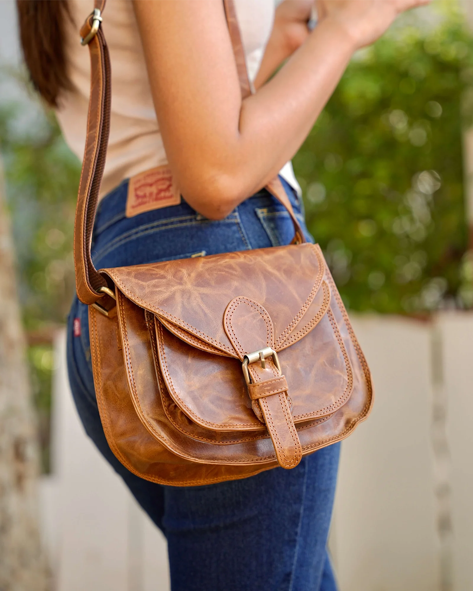 A woman wearing a beige top and blue jeans carries a brown distressed real leather crossbody bag with a flap closure and gold-tone buckle. 100% genuine full grain leather, natural patina