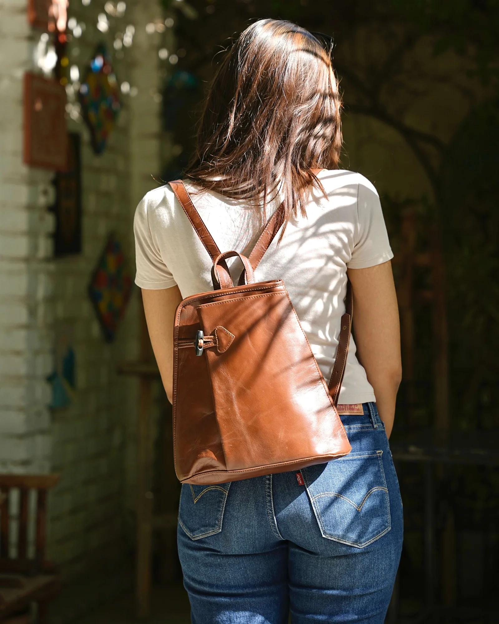 A woman viewed from behind wearing a brown leather backpack with a front buckle strap, in an outdoor setting with dappled sunlight.