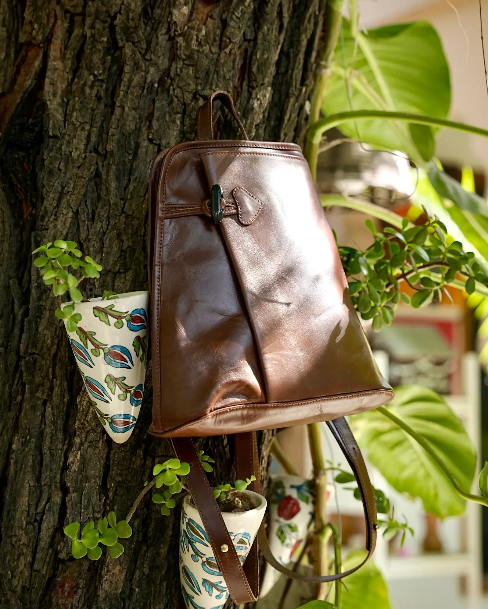 A brown leather backpack with a front flap and buckle closure hangs on a tree trunk, surrounded by green plants and small hanging pots.