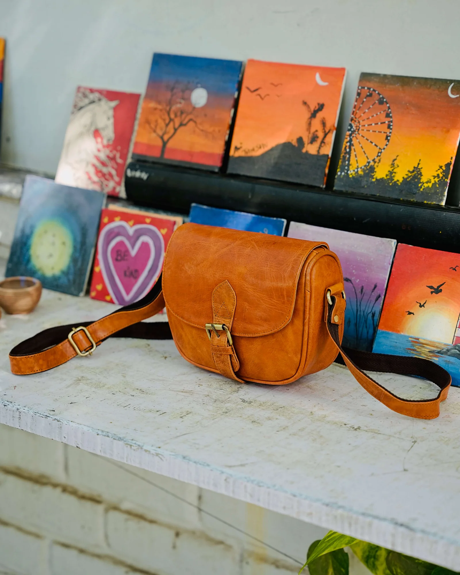 A tan real leather crossbody bag with a buckle closure and adjustable strap, displayed on a white wooden surface in front of a row of colorful painted canvases. 100% genuine full grain leather, natural patina