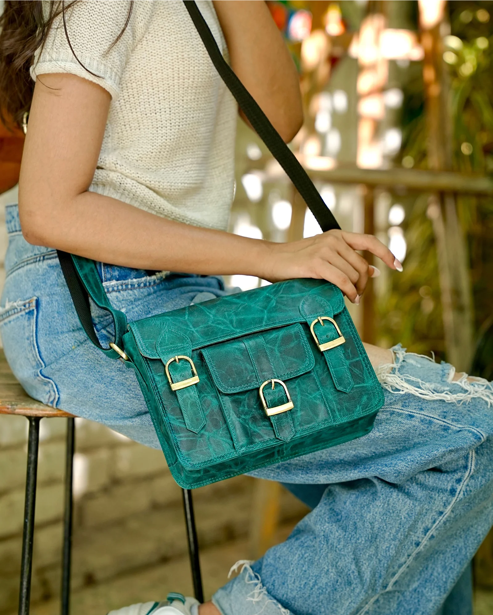 A woman wearing a white knit top and distressed blue jeans sits on a stool, holding a teal crocodile-embossed crossbody bag with gold-tone hardware and a black strap.