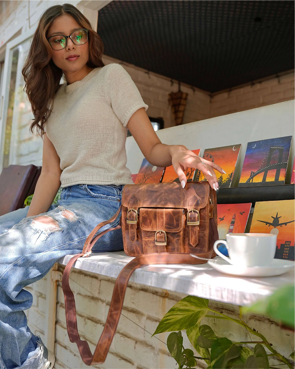 A woman wearing a cream knit top and ripped blue jeans sits on a white brick ledge, holding a distressed brown leather crossbody bag with gold-tone hardware. A white coffee cup sits on a saucer next to the bag, with framed artwork and green leaves in the background.