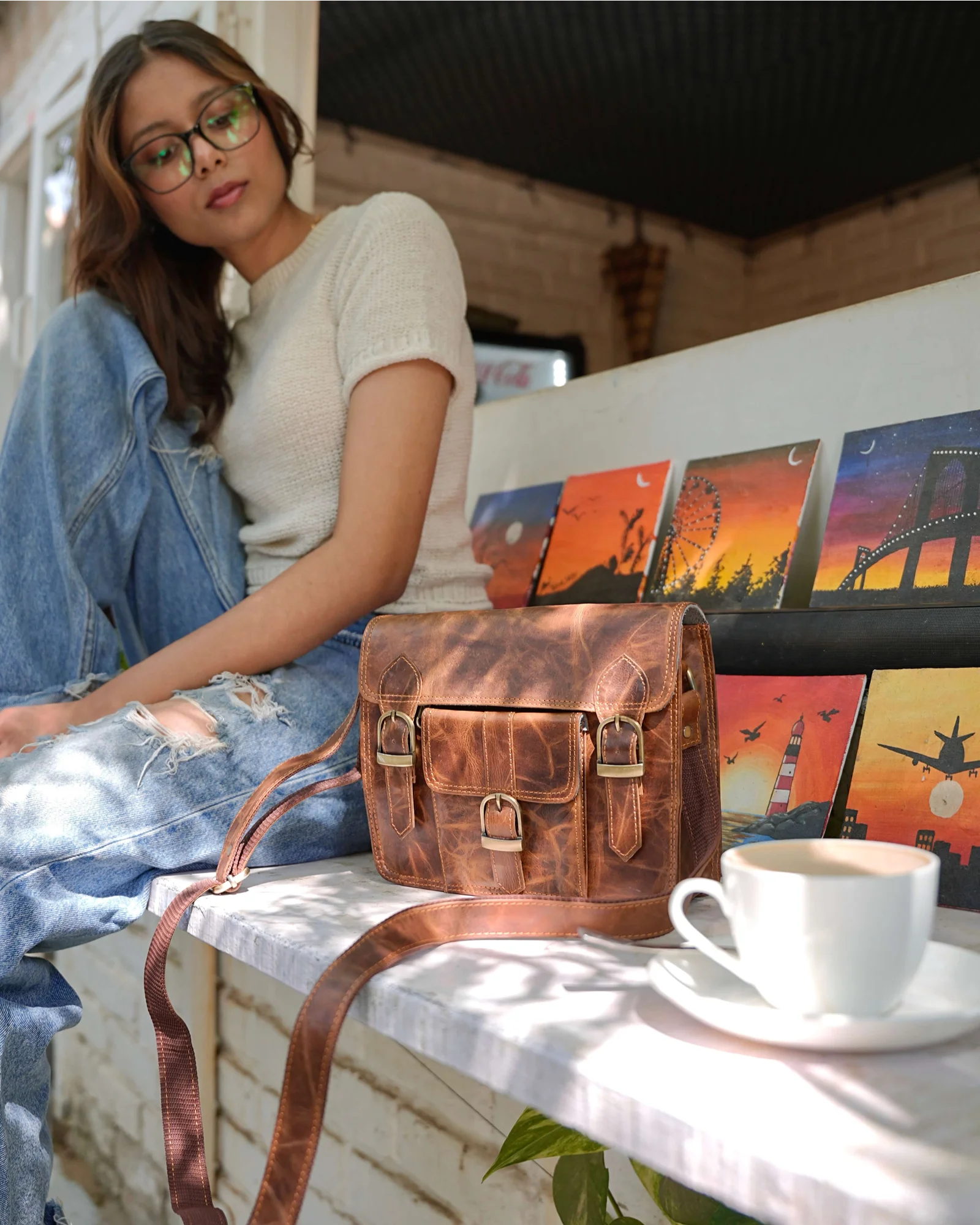 A woman wearing glasses and a cream t-shirt sits on a bench in a cafe setting, with a distressed brown leather crossbody bag placed on the bench in front of her. A white coffee cup is on a saucer to the right, and colorful art prints are visible in the background.