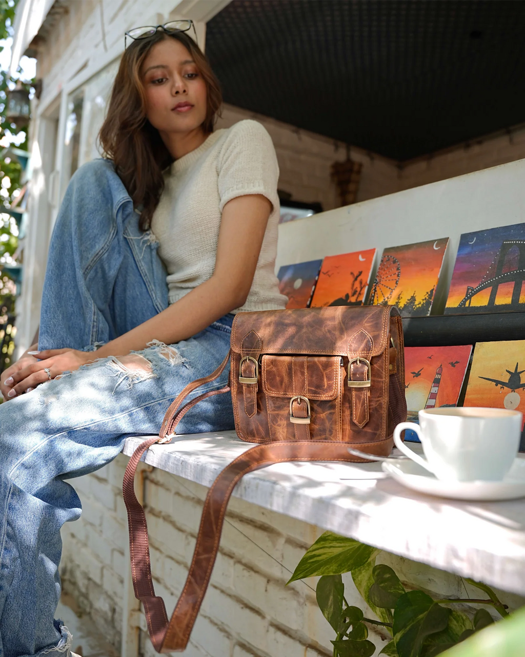 A woman sits on a porch ledge wearing a cream ribbed top and distressed blue jeans, with a brown distressed leather satchel bag resting on the ledge in front of her. The bag has brass hardware and a long strap.
