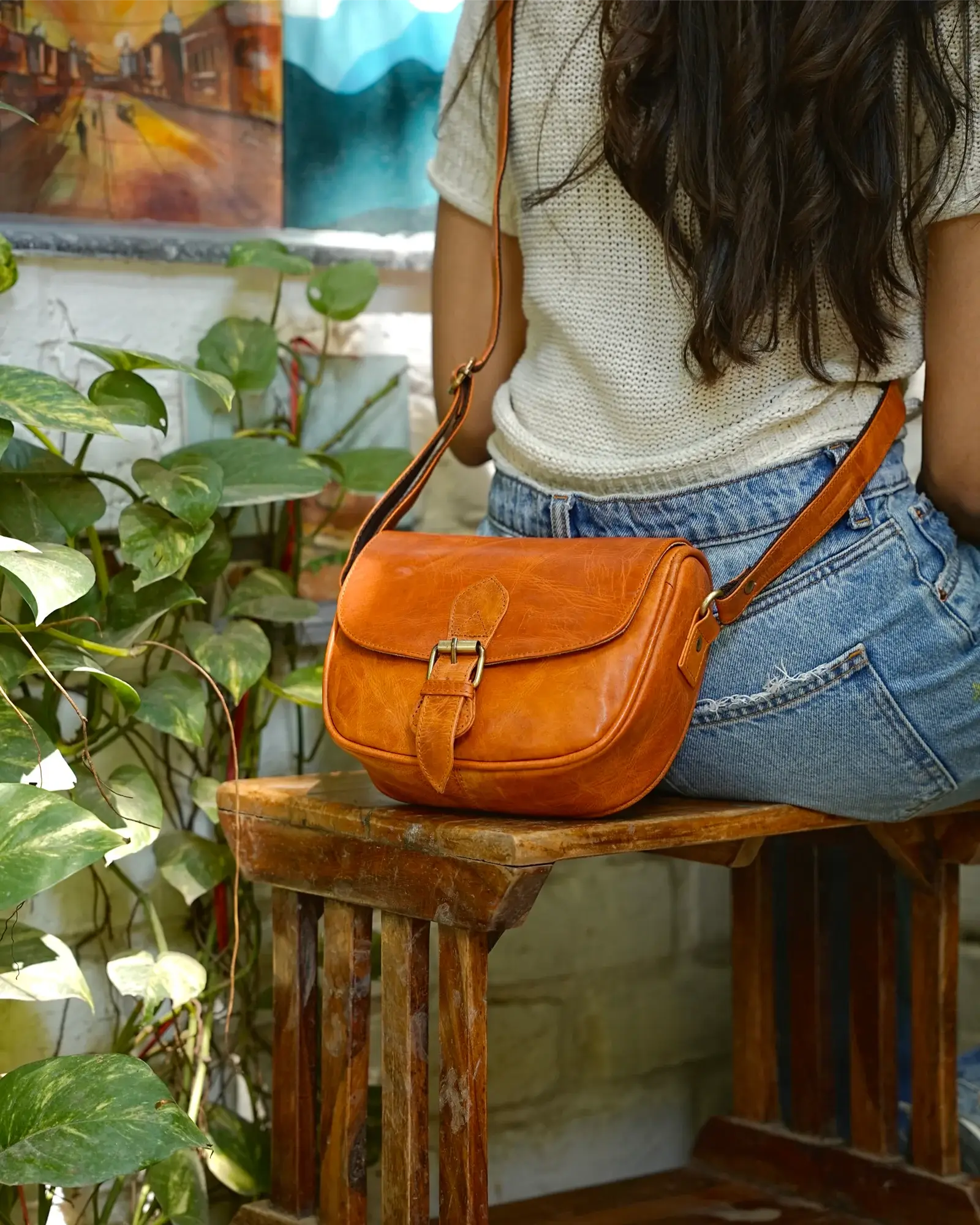 A woman wearing a cream knit top and blue jeans sits on a rustic wooden bench, viewed from behind, with a warm brown leather crossbody bag resting on the bench in front of her. 100% genuine full grain leather, natural patina