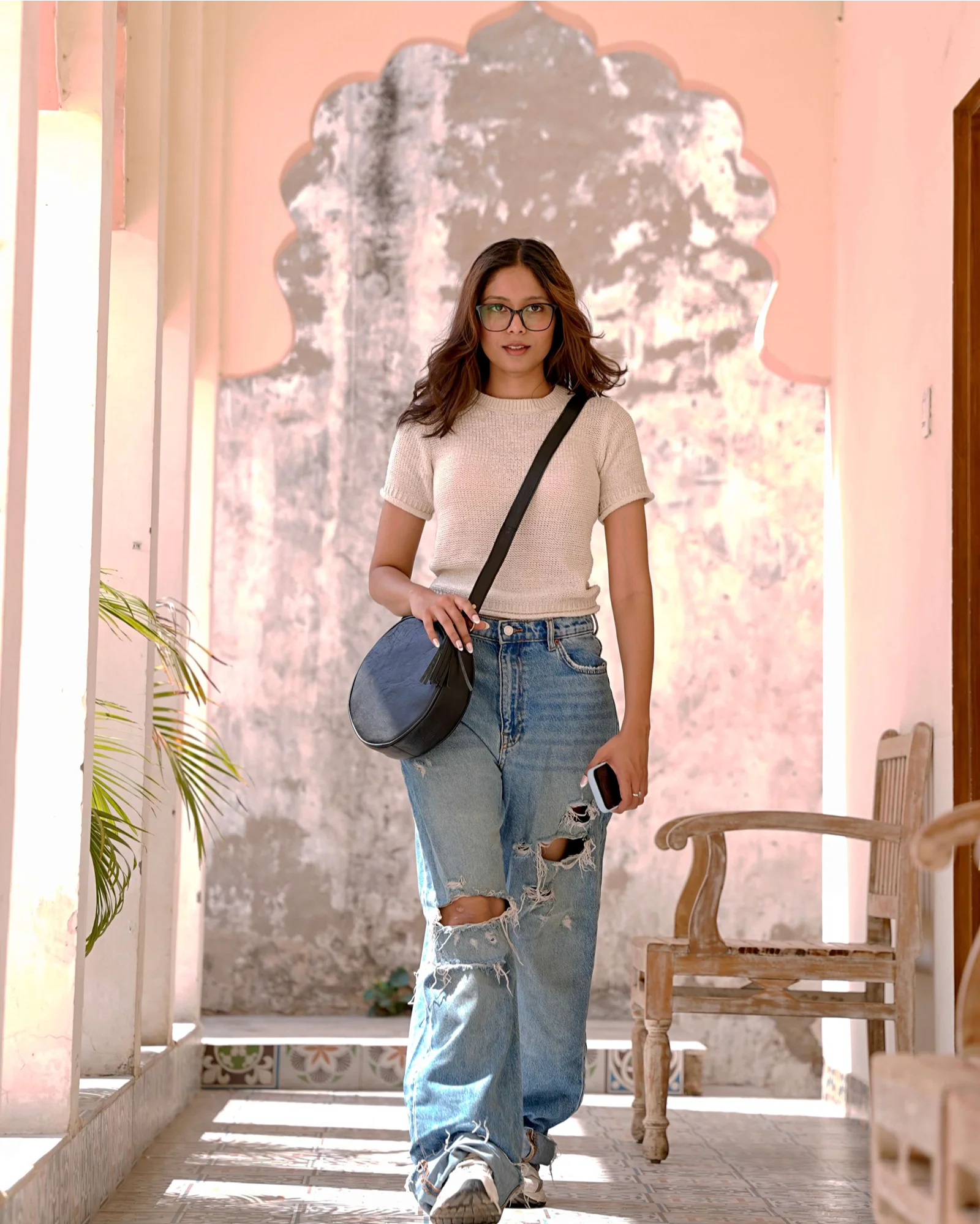 A woman wearing a beige t-shirt, ripped light-wash blue jeans, and glasses walks down a sunlit corridor with a pink wall and arched doorway, carrying a dark blue crescent-shaped crossbody bag.