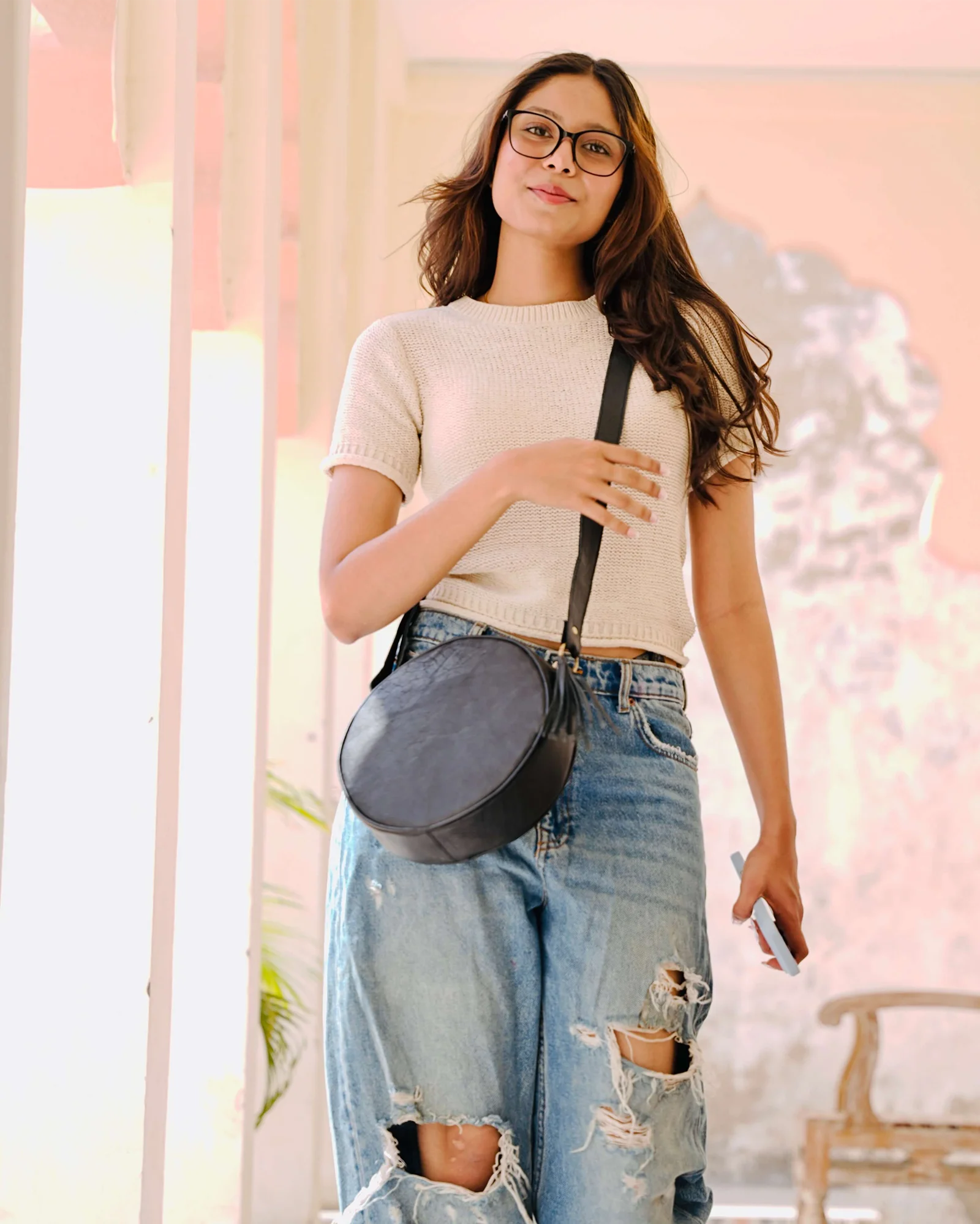 A woman wearing round eyeglasses, a beige short-sleeve knit top, and distressed light-wash blue jeans, carrying a black round crossbody bag with a thin black strap.