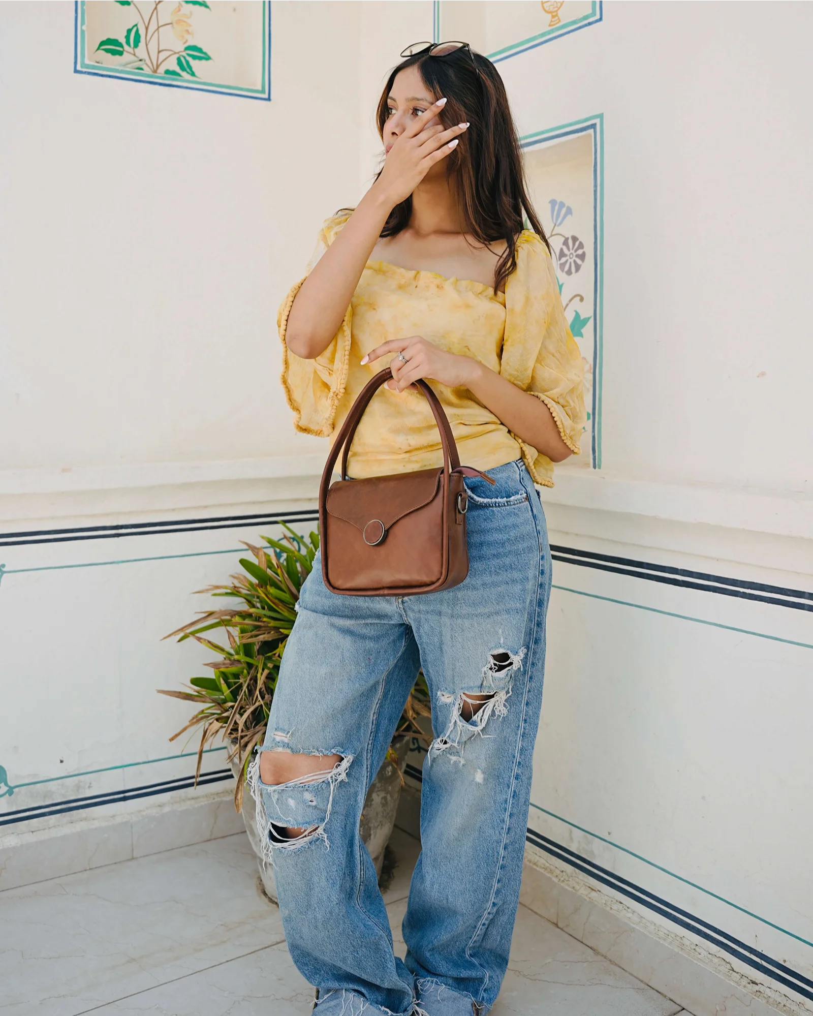 A woman wearing a yellow ruffled off-the-shoulder top, distressed light blue jeans, and holding a brown leather handbag, standing against a white wall with decorative blue lines. 100% genuine full grain leather, natural patina