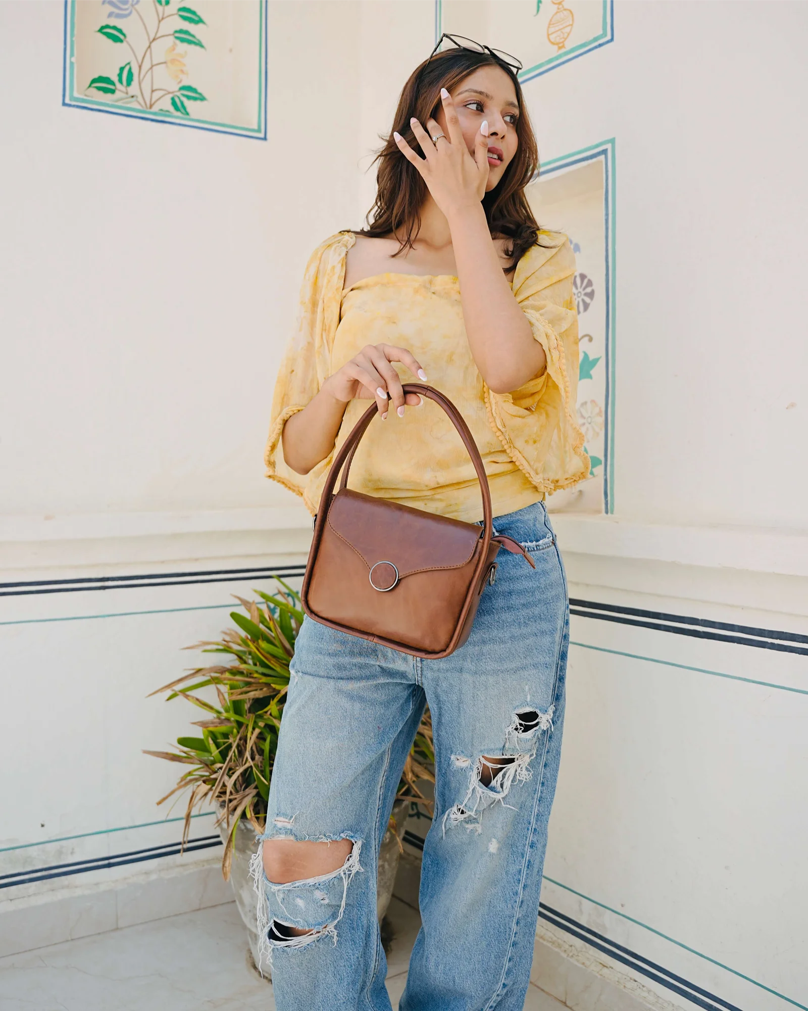 A woman wearing a yellow ruffled top and ripped blue jeans holds a brown leather handbag with a top handle, standing against a white wall with decorative panels.