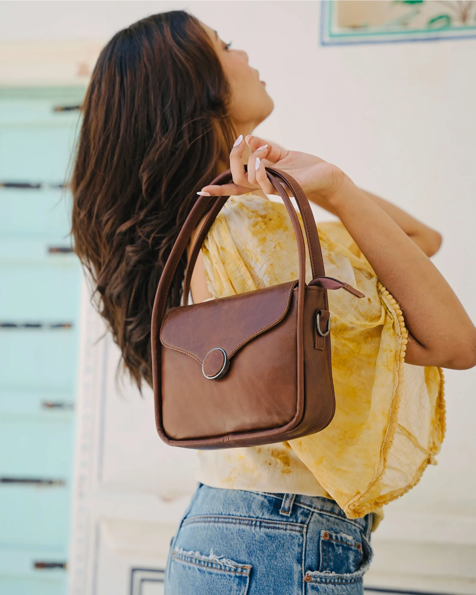 A woman in a yellow top and blue jeans holds up a brown leather shoulder bag with a top handle and a circular metal clasp, set against a light-colored background.