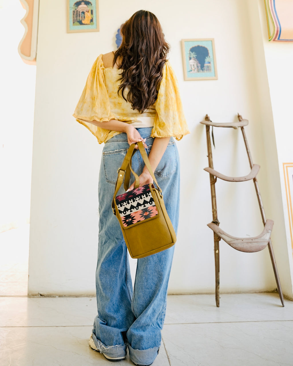 camel crossbody bag with patterned flap held by a person wearing blue jeans.