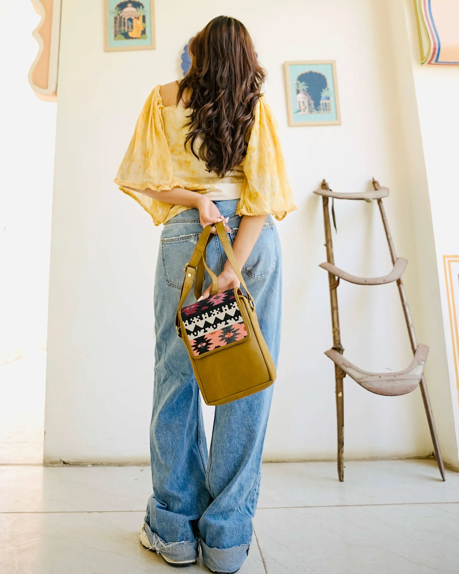 A woman stands with her back to the camera wearing a yellow off-the-shoulder blouse, wide-leg blue jeans, and holding a mustard yellow handbag with a colorful patterned front pocket, in a brightly lit room with framed art and a wooden stool.