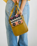 camel crossbody bag with patterned flap held by a person wearing blue jeans.