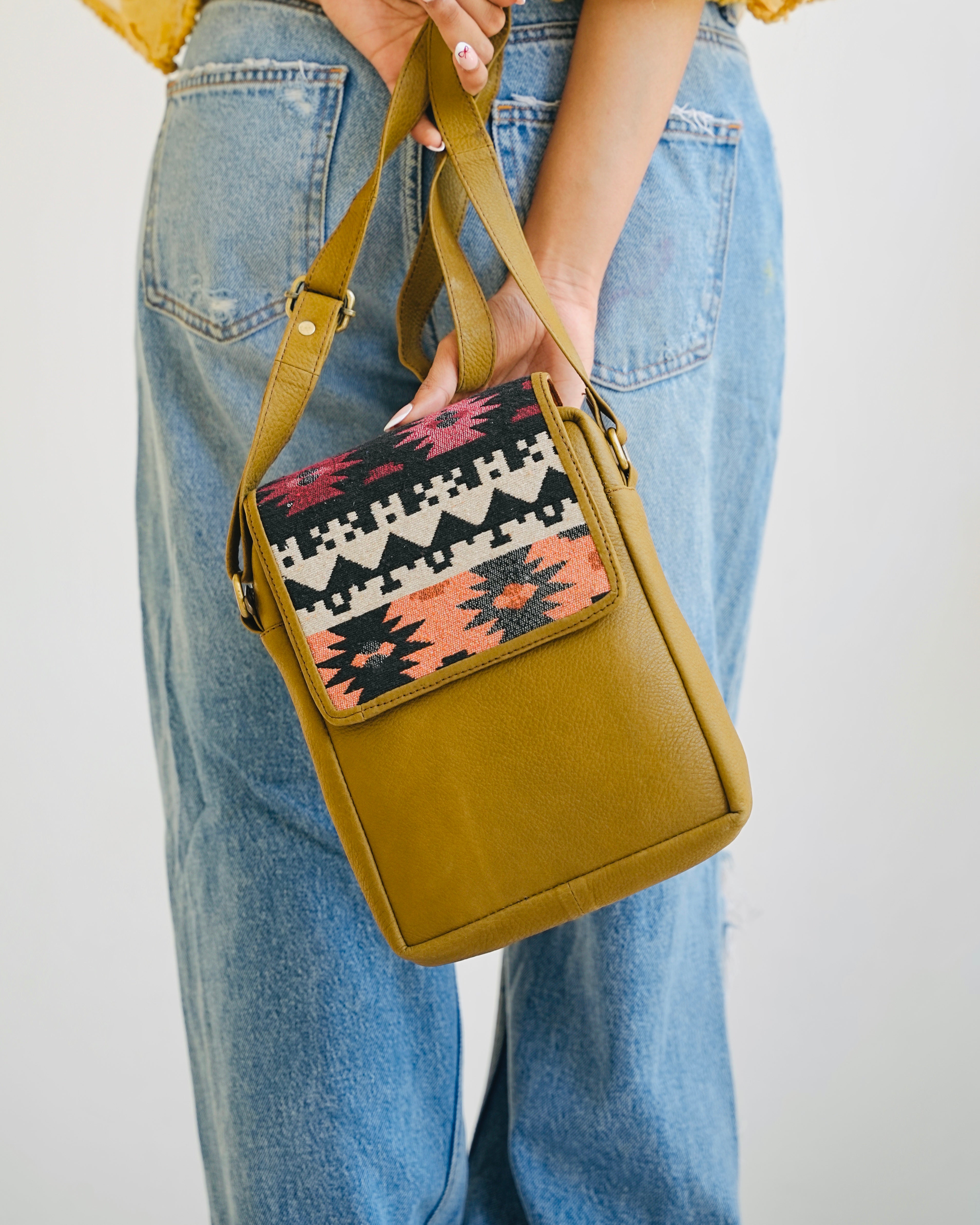 camel crossbody bag with patterned flap held by a person wearing blue jeans.