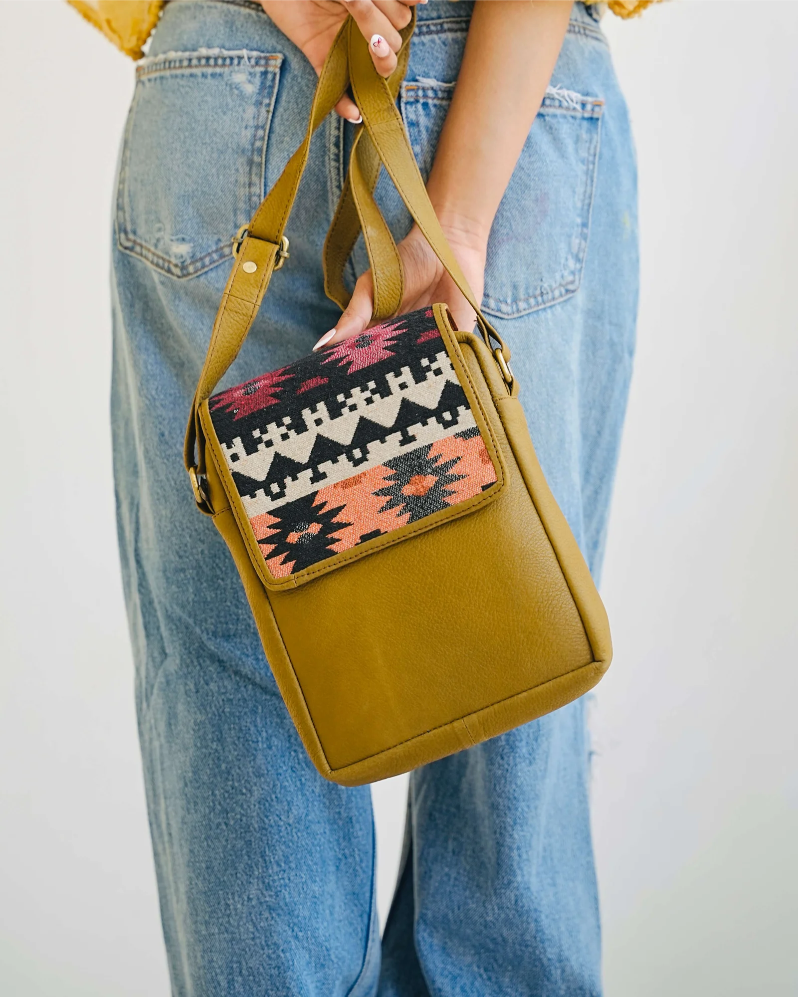 A person wearing blue jeans holds a mustard yellow real leather crossbody bag with a colorful geometric patterned front flap. The bag has tan straps and a solid mustard yellow body, shown against a plain light background. 100% genuine full grain leather, natural patina
