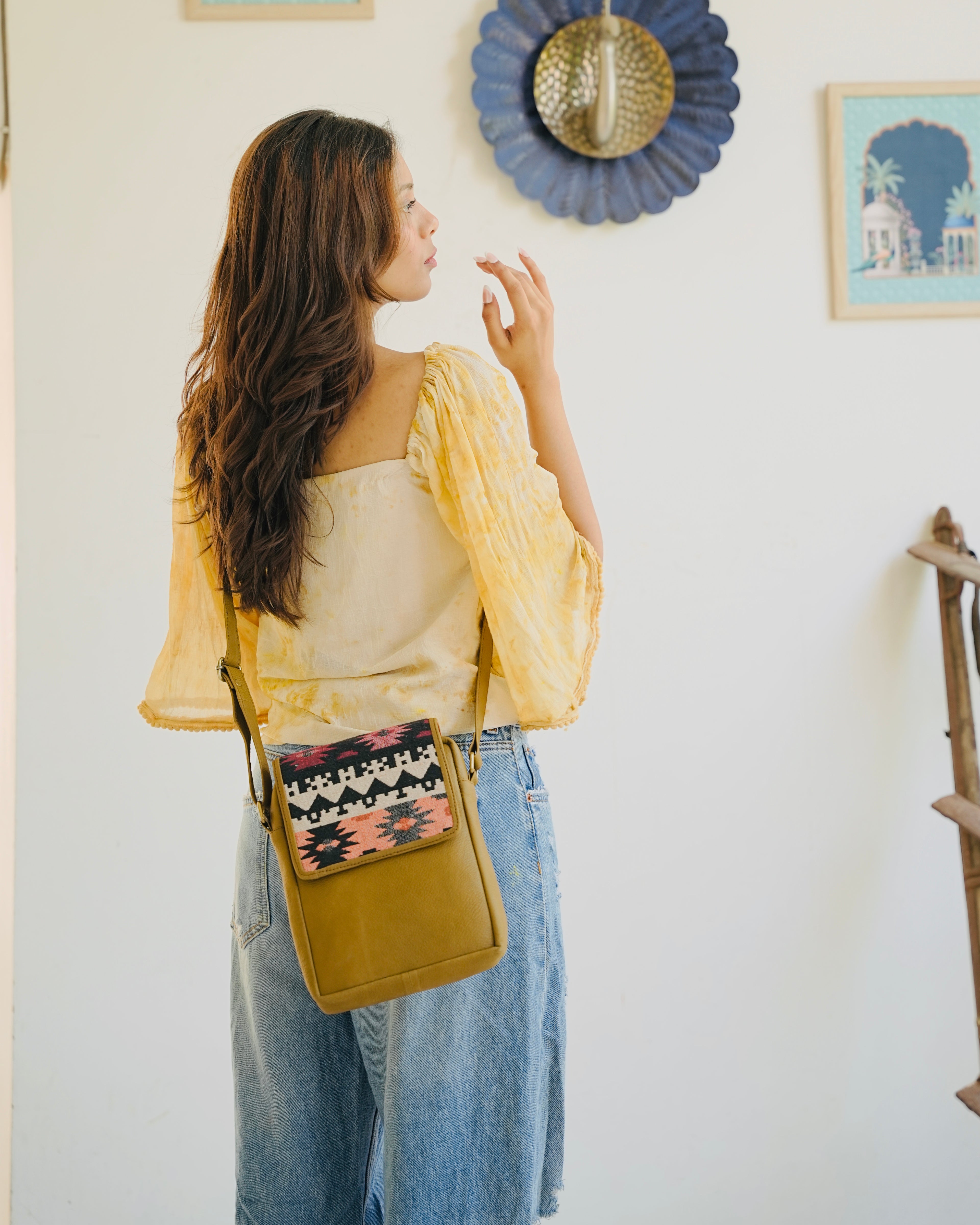 camel crossbody bag with patterned flap held by a person wearing blue jeans.