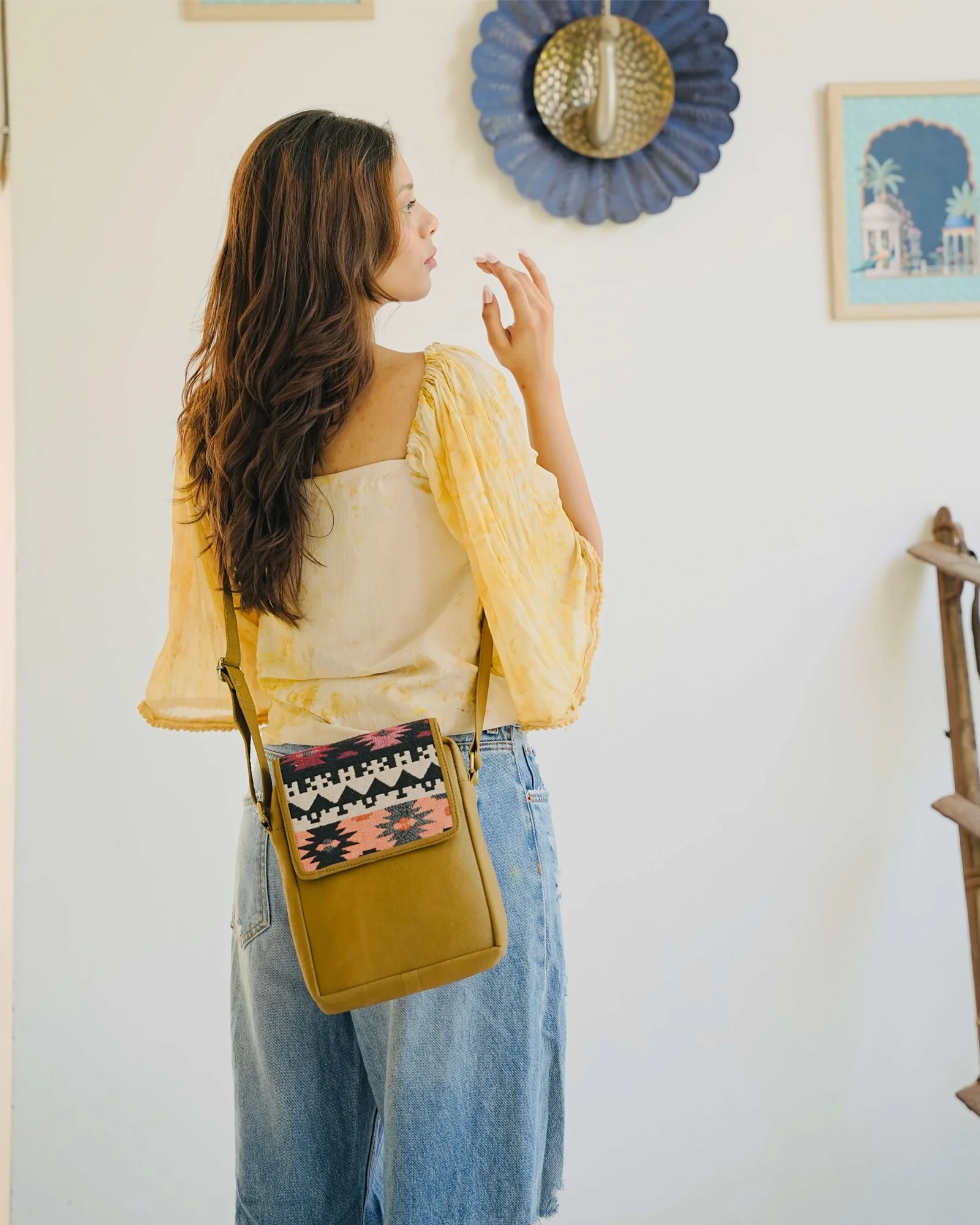 Woman wearing a yellow top and blue jeans with a patterned bag, standing indoors.