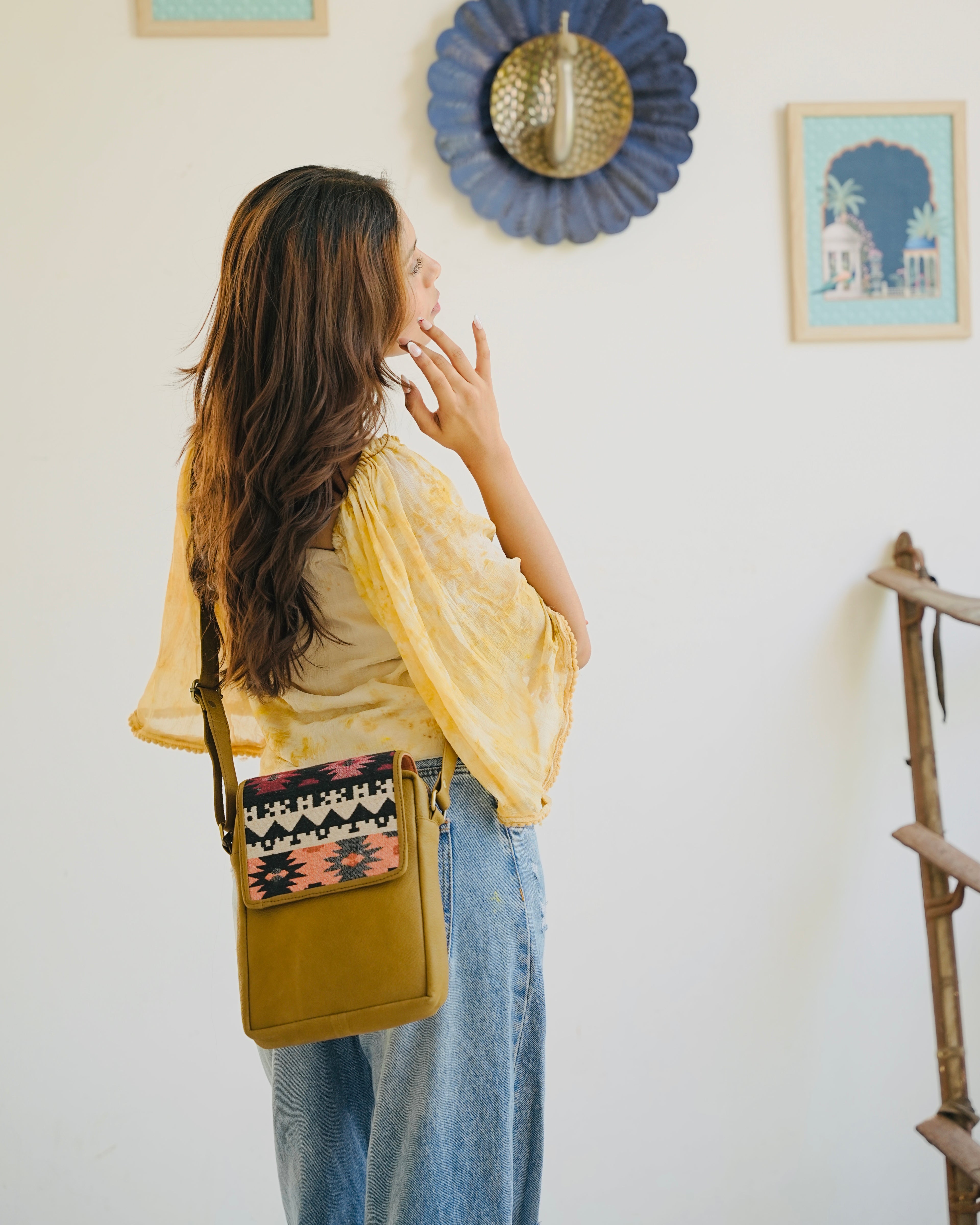 camel crossbody bag with patterned flap held by a person wearing blue jeans.