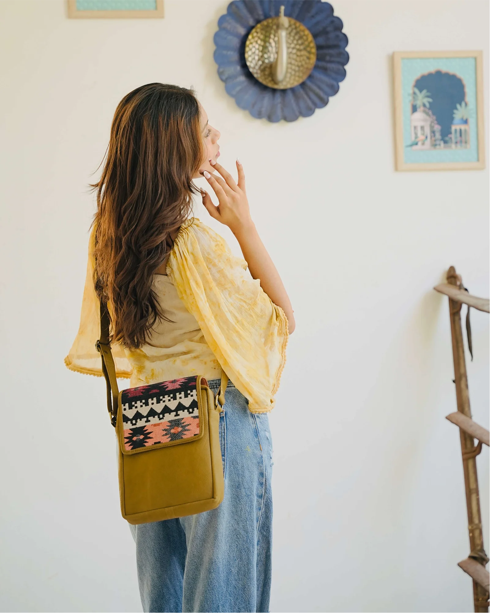 A woman with long brown hair is shown from behind wearing a yellow top and blue jeans, carrying a mustard yellow crossbody bag with a colorful patterned flap. She is standing in a brightly lit room with a white wall decorated with framed art and a blue flower-shaped wall hanging.