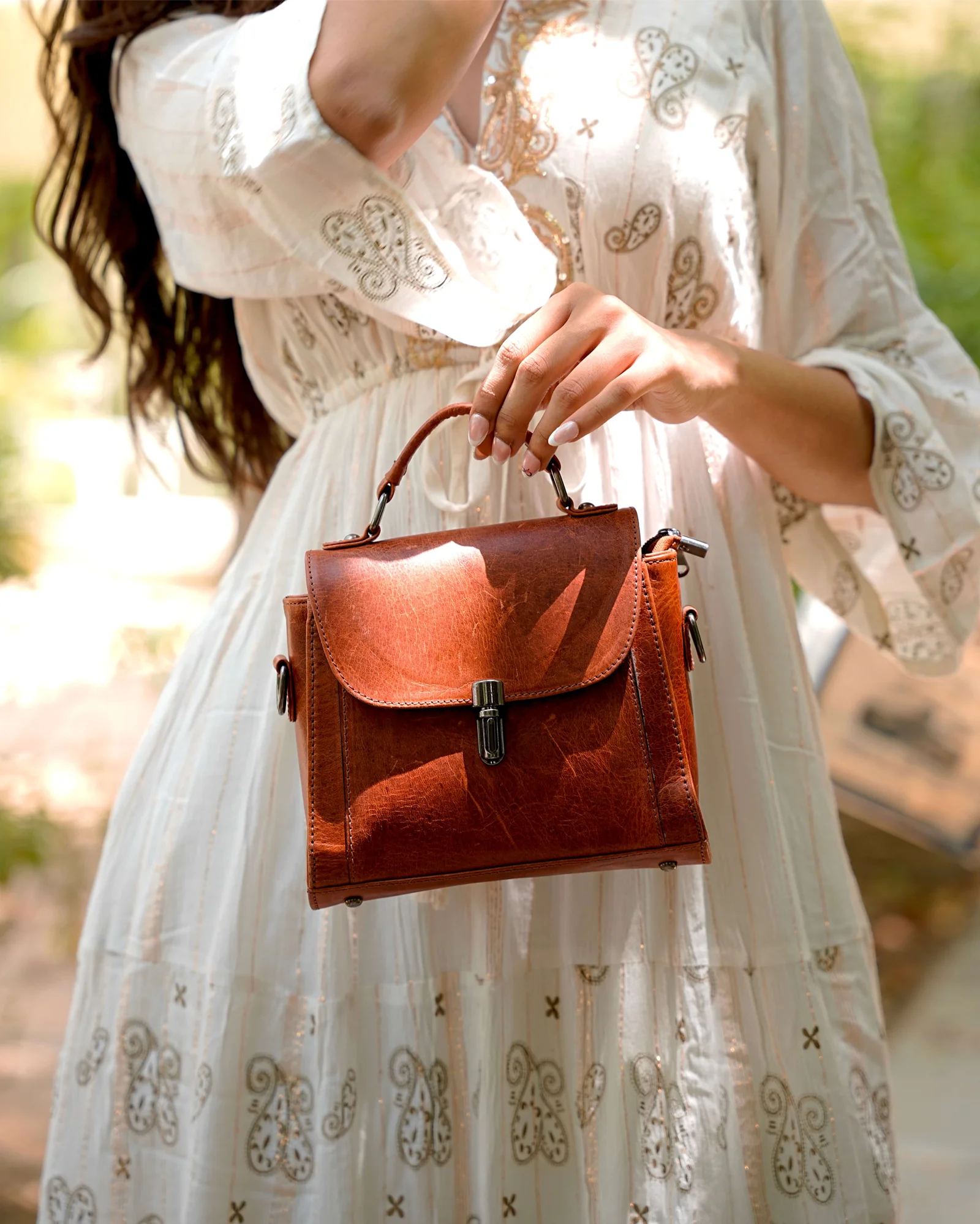 A woman in a white embroidered dress holds a small brown leather top-handle bag with a front flap and metal clasp in an outdoor setting.