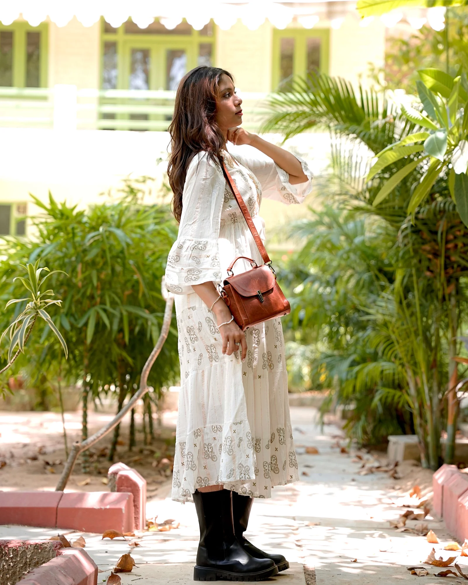 A woman stands outdoors in a garden setting wearing a white embroidered midi dress, a brown leather crossbody bag, and black rubber boots.