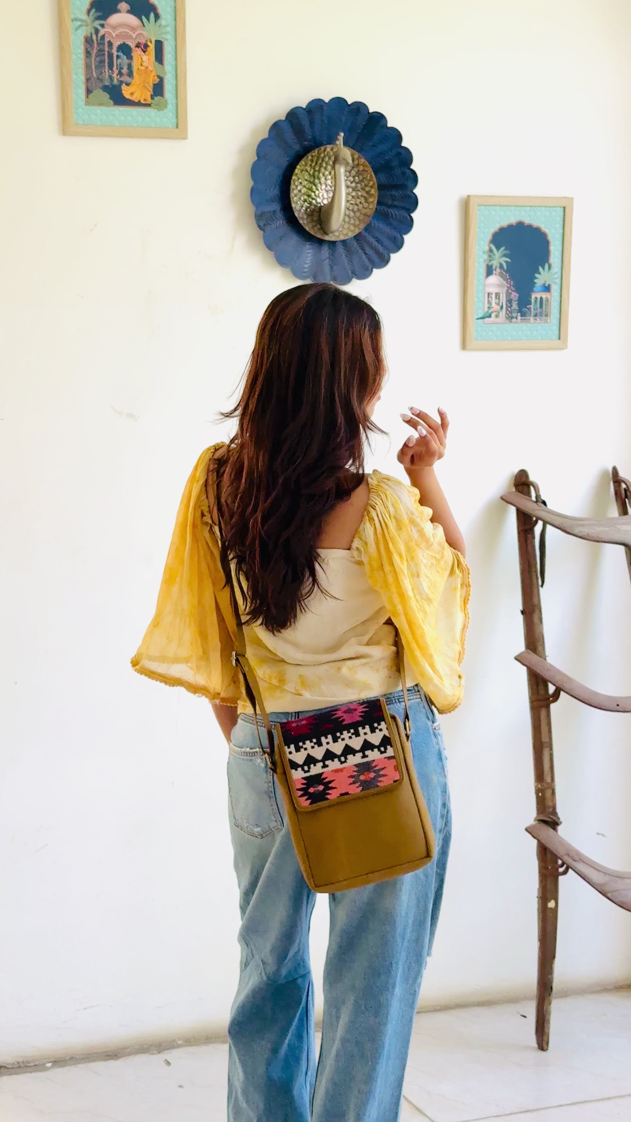 camel crossbody bag with patterned flap held by a person wearing blue jeans.